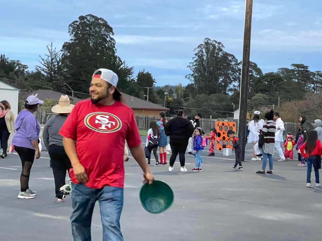 Outdoor community event: A man wearing a San Francisco 49ers shirt carries a green bucket during an outdoor church community event with families and children in the background.